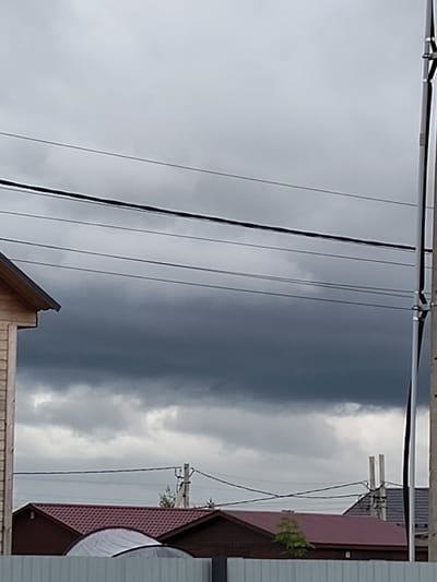 The image depicts an outdoor scene with a cloudy sky. Several power lines are visible, stretched across the frame. In the lower part of the image, the roofs of houses with dark red tile are visible, along with a section of a light-colored fence in the foreground. A portion of a wooden building is visible on the left edge. The lighting suggests an overcast day.