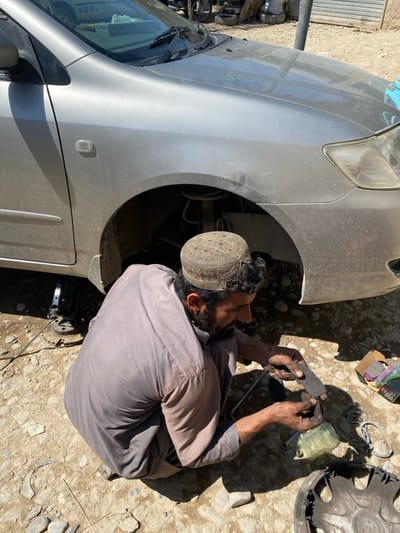 The scene is an outdoor vehicle repair area in کوکران, Afghanistan. A man, consistent with Afghan ethnicity, with a dark beard and mustache, wearing a patterned round cap and a light brown long-sleeved tunic, squats on unpaved dirt and gravel. His hands are covered in grease. He holds and examines a dark, rectangular brake pad. Immediately to his right, a silver sedan is visible; its front right wheel is removed, and the vehicle is elevated by a black hydraulic jack positioned under the frame. Exposed within the wheel well are the vehicle's suspension components. On the ground near the man are an open cardboard box containing new brake pads, a damaged dark grey hubcap, various small metal tools, and a clear plastic bag. The environment is dusty, illuminated by bright daylight, with distant background elements including other vehicles and rudimentary structures. On the car's dashboard, visible through the windshield, is a white box labeled "ALKOZAY".