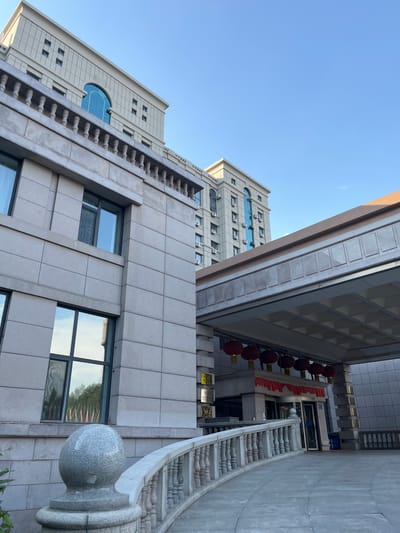 The image shows the exterior of a building, possibly a hotel or a large office building, in Changchun City, China. The architecture is modern with classical influences, featuring light-colored stone facades, arched windows, and ornate railings. In the foreground, a curved stone pathway leads towards an entrance sheltered by a covered walkway. Red lanterns and festive decorations hang from the walkway, suggesting a celebratory occasion. The sky is clear and blue, indicating daytime. The buildings are tall and imposing, with numerous windows and air conditioning units visible. There are no people clearly visible in the scene. The overall impression is one of a grand and well-maintained establishment.