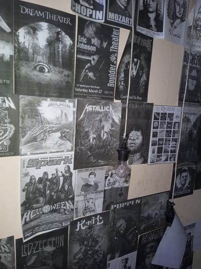 A collage of black and white posters covers a wall, showcasing various music-related content. Featured are bands like Dream Theater, Metallica, Queen, Led Zeppelin, and Helloween. There's also a poster for Eric Johnson at the Boulder Theater. Classical music composers Chopin, Mozart, Bach, Tchaikovsky, and Vivaldi are also represented. The image includes rock magazine covers and posters of lesser-known bands and musicians. An exposed light bulb hangs in front of the posters, casting light and shadows on the display.