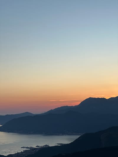This is a vertically oriented landscape photograph taken at sunset. The image features a vast sky transitioning from a pale blue to a warm orange and pink hue, indicative of the golden hour. Silhouetted against this vibrant sky are the dark, layered shapes of mountains.
Below the mountains, a body of water, likely a bay or lake, reflects the soft light of the sky. Along the shoreline, a town or village is visible, with lights beginning to twinkle as dusk settles. Several boats and ships are moored in the water, adding a sense of human presence to the scene.
The overall mood is serene and tranquil, capturing the quiet beauty of a coastal landscape at the end of the day. The location is Žanjev Do, Montenegro. There is no visible text in the image.