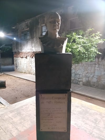 The image is a nighttime photograph of a bust of a woman on a stone pedestal in front of a building. The bust is made of a light-colored material, possibly stone or concrete, and depicts a woman with short hair and a stern expression. She is wearing a draped garment around her neck and shoulders. The pedestal is made of dark stone and has a plaque on the front with an inscription in Spanish. The inscription appears to be a dedication to "Petronila Concepción de Mata Romero."
Behind the bust and pedestal is a two-story building with a weathered facade and barred windows. To the right of the building is a dense cluster of green foliage. The ground in front of the pedestal is covered in dirt and a paved walkway. The lighting suggests it is an outdoor public space, likely a park or plaza. The scene appears to be in Santa Ana, Venezuela, as indicated by the inscription on the plaque.