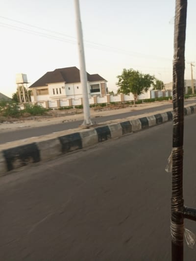 The image shows a street scene in Maiduguri, Nigeria. The image was likely taken from a moving vehicle, as the street appears blurred. The scene is a residential area with a large, two-story house on the right side of the road. The house has a white exterior with a brown roof. There is a tall, white pole with electrical wires running across the street in front of the house. There is a water tower on the left side of the road in front of the house. There is a single tree in the middle ground with green foliage. The ground is dusty and brown, and there is a concrete curb on the edge of the road. The street has black and white markings. The image is taken in the late afternoon or early evening, as the sky is a light blue color. The image is taken from a point of view that is close to the ground and looking straight ahead. The image is taken from a moving vehicle. The black pole in the foreground is blurred and distorted, as the vehicle was moving when the image was captured. The image is out of focus in general due to the motion of the vehicle.