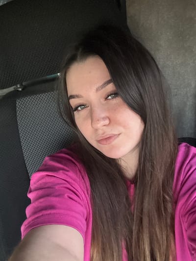 A young woman with long, dark brown hair is taking a selfie in what appears to be the interior of a vehicle. She is wearing a bright pink t-shirt. Her head is tilted slightly, and she is looking directly at the camera. The background shows the textured upholstery of a car seat and a portion of the car's interior, suggesting the location could be within Krasnokamsk, Russia, though this is speculative without further information. The lighting is consistent with indoor, possibly natural light, and the focus is on the woman's face.
