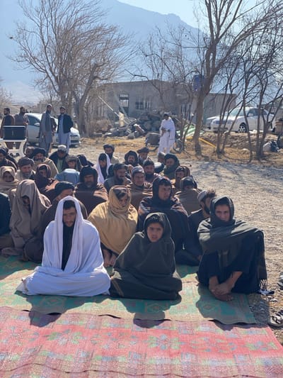 A group of men sit cross-legged on rugs in an outdoor setting. Most are wrapped in shawls or blankets, predominantly in shades of brown, white, and black. Some men are looking directly at the camera. Behind them, there is a building, trees, and a mountainous landscape. A few men stand in the background, dressed in formal attire. The light suggests it is daytime.