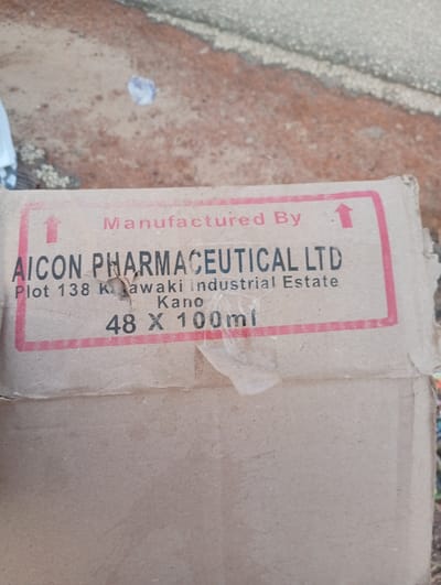 The image shows a cardboard box with printing. The text on the box says "Manufactured By AICON PHARMACEUTICAL LTD Plot 138 Kwaki industrial Estate Kano 48 X 100ml". The box is damaged with a tear in the middle and tape on the bottom right. The box is lying on the ground, which appears to be dirt with some small stones and debris. There is a white plastic bag in the background. The scene is outdoors. The city of Gulak, Nigeria is not visible in this image.