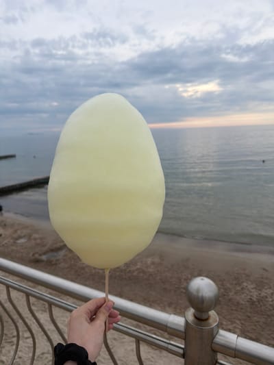 A person's hand holds a pale yellow cotton candy stick in front of a beach scene in Zelenogradsk, Russia. The cotton candy is the primary subject, obscuring the background partially. The hand has a black scrunchie on the wrist. The background shows a sandy beach, a metal railing, and the sea under a cloudy sky. The overall atmosphere is calm and slightly overcast.