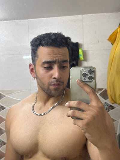A man is captured in a mirror selfie, shirtless and displaying a muscular physique. He appears to be in his 20s or 30s, with dark, curly hair and neatly trimmed facial hair, including a beard or heavy stubble. He has a silver chain around his neck. His expression is relatively neutral, with a slight furrow in his brow, giving a contemplative or serious look.

He holds a smartphone, likely an iPhone (suggested by the triple-lens camera module and flash on the rear), in his right hand, positioned to capture his reflection in the mirror. The phone is encased in a clear or light-colored protective cover.

The setting is an indoor space with light-colored, possibly tiled walls, which could indicate a bathroom or changing area. In the upper part of the reflection, a vibrant yellow fabric or garment is visible, possibly hanging or draped. Further in the background, out of focus, are faint dark objects and a small container with green and yellow accents on a surface. No discernible text is visible on the phone screen or in the background.