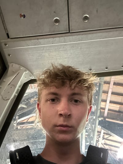 The image displays a close-up selfie of a young male inside a metallic lift cabin. The subject is a Caucasian male, approximately 18-25 years old, with light, disheveled hair and a neutral expression, looking directly forward. He is wearing a dark-colored shirt and black protective shoulder pads, one bearing the text "BONC" and the other "PRO". The cabin interior is constructed from light grey metal panels secured with visible rivets; two circular indentations and a small, multicolored circular sticker are present on the upper panels. Through the window to the subject's left, the exterior reveals a large, intricate structure composed of numerous metal beams and supports, indicative of a gondola or telecabin lift station. The location is an operational lift system in Les Gets, France.