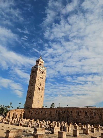 The image displays a prominent architectural landmark under a bright blue sky. The main subject is a tall, square-based minaret constructed from light brown or sandy-colored stone. The minaret features intricate geometric patterns and decorative elements carved into its surface, characteristic of Almohad architecture. Its spire tapers to a point, topped with what appear to be three golden or brass spheres of decreasing size, culminating in a decorative finial.
To the right of the minaret, a lower, longer building or wall extends horizontally across the frame. This structure is also made of similar light-colored stone and exhibits a series of recessed arched openings along its facade, some of which appear to be windows or decorative niches. In the foreground, below this longer building, there is an area filled with numerous short, rectangular stone structures or foundations, suggesting either ruins, a garden layout, or an unfinished construction.
Sparse vegetation, primarily palm trees, can be seen behind the lower building and near the base of the minaret, adding a touch of greenery to the arid-looking landscape.
The sky is a dominant feature, occupying the upper and left portions of the image. It is a vibrant, clear blue, adorned with scattered, wispy white clouds that stretch across the expanse, creating a sense of depth and movement.
The photograph appears to have been taken during the daytime, likely late morning or early afternoon, given the bright, even illumination and the relatively high position of the sun, suggested by the subtle shadows cast by the structures. The overall impression is one of historical grandeur and architectural beauty against a natural, serene backdrop. No discernible text is visible in the image.
