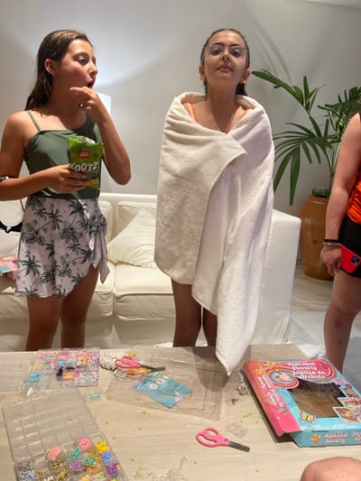 The image shows an indoor scene. Two females are visible. One is eating from a bag of "KOOTZ" brand food product. She is wearing a green top and a skirt with a palm tree pattern. The second female is draped in a white towel and looking toward the camera. Other objects include a box labeled "Alphabet Jewelry Joyeria de Afabeto," a plastic container with beads, a pair of pink scissors, and a white sofa. The image is rotated counter-clockwise. The location is Marina Maroma, Mexico.