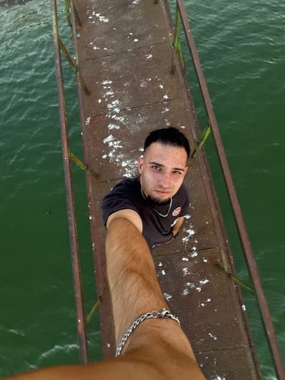A man is taking a selfie from a high vantage point, looking down at the camera. He is on a rusted metal pier that extends over green water. The pier is speckled with white, likely bird droppings. The man has dark hair, a short beard, and is wearing a dark t-shirt with a silver chain necklace and bracelet. The water appears to be a reservoir or a wide river. The image does not provide enough information to determine the specific location within the city of Shumen, Bulgaria.
