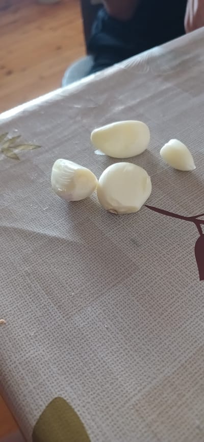 Five peeled garlic cloves are scattered on a light-colored, textured tablecloth. The tablecloth has a faint floral pattern. In the background, a person's dark clothing is visible, and beyond that, a wooden floor. The scene appears to be indoors.
