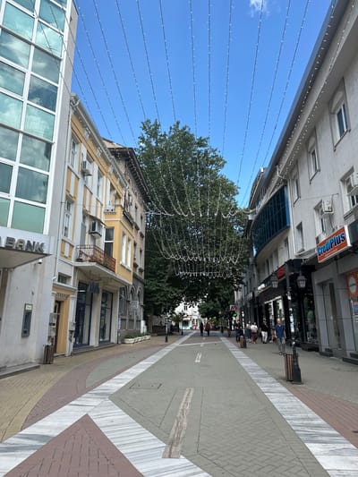 The image captures a street scene in Haskovo, Bulgaria, likely during daylight hours under a clear blue sky. The perspective is from a pedestrian's viewpoint, looking down a wide, pedestrianized street lined with buildings on both sides. A large, mature tree is centrally located in the distance, its dense foliage partially obscuring the view beyond.
Suspended above the street are numerous strands of small white lights, forming decorative arches or canopies, suggestive of festive decoration or street lighting. The buildings flanking the street exhibit a mix of architectural styles, with some featuring classical facades and others more modern designs.
On the left, a modern building with a glass facade and visible internal blinds is prominent, alongside an older building with yellow and white striped detailing, featuring several windows and balconies. A sign reading "BANK" is visible on the modern building.
On the right, buildings are painted in lighter colors, with visible windows and signage. One sign reads "CrediPlus" with Bulgarian text beneath it. Street lamps and some potted plants are also visible along the sidewalks.
The street itself is paved with a combination of materials, including grey cobblestones and decorative sections of white and reddish-brown marble or stone in geometric patterns. Several individuals are walking along the street in both directions, appearing to be engaged in everyday activities. Some are walking alone, while others are in small groups. The overall atmosphere appears calm and urban.