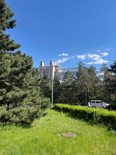 The image shows a grassy area adjacent to trees and a building complex in Changchun City, China. The sky is bright blue with scattered clouds. A white vehicle is parked near a hedge on the right side of the frame. A light post is visible amidst the trees. The building is in the background.