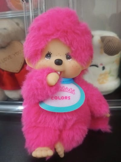 A pink Monchhichi doll stands centered. It has furry pink hair and body, a beige face with freckles, a black nose, and large, reflective eyes. The doll wears a white bib with "Monchhichi COLORS" printed in pink and outlined in teal. A pacifier is near its mouth. Blurred background figures include a teddy bear with a heart and a plush animal. The doll is positioned on a dark surface.