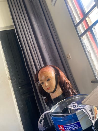 A mannequin head wearing a wig sits in front of a dark grey curtain and a dark wooden door in what appears to be a room in Kubwa, Nigeria. To the right of the mannequin, a window with a black metal grate reveals a glimpse of the outdoors, with some peeling paint on the window frame. Below the mannequin's head, a blue metal tin labeled "Premium Quality Cookies" is visible, with a black and white striped ribbon peeking out from inside it. The mannequin has light brown hair styled in waves, and is positioned at an angle to the camera. The lighting in the room suggests it is daytime.