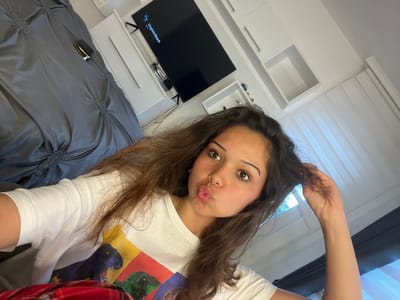 A young woman with long brown hair and large brown eyes is taking a selfie in a bedroom. She is wearing a white t-shirt with a colorful dinosaur print. Her lips are puckered as if she is blowing a kiss. Her right hand is raised and pulling on her hair. In the background, a large television screen displays the words "Virginia Beach" and a silhouette of a dog. The room appears to be a bedroom, with a bed covered in a gray duvet on the left side of the image, and white cabinets and shelves on the right. The overall lighting suggests it is daytime. The location of Zlatograd, Bulgaria is not discernible from the image.