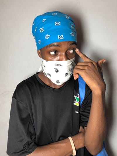 A single individual is depicted against a plain, light grey background in Ngleshi Amanfro, Ghana. The person, with dark skin, is seen from the chest up, angled slightly to the left. They are wearing a bright blue bandana covering their head and a white bandana with black paisley patterns functioning as a face mask, obscuring the nose and mouth. A black, short-sleeved t-shirt with a multi-colored logo and "CAN" text on the left chest is worn. A light-colored bracelet is present on the right wrist. A blue strap from a bag is visible over the individual's right shoulder. The person's right hand is raised, with the index finger touching the right temple/eyebrow area, while the eyes are directed to the viewer's left.