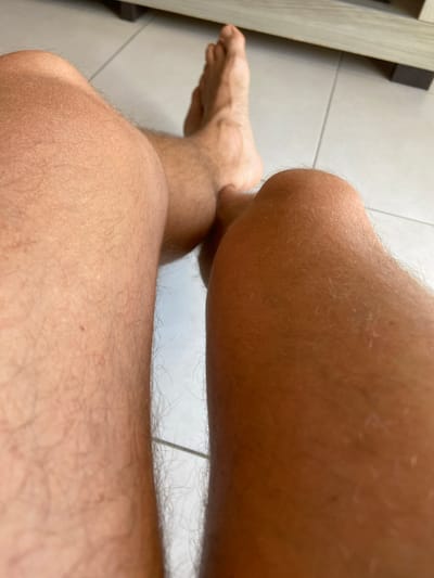 The image shows a close-up view of a person's legs, including their knees and feet. The person appears to be sitting on a tiled floor, with the legs angled towards the viewer. The lighting suggests an indoor setting, possibly a home or hotel room. The background includes a glimpse of a piece of furniture, potentially a bed frame or cabinet. The overall impression is one of relaxation or casual repose. Without further context, it is not possible to determine the specific location within João Pessoa, Brazil.