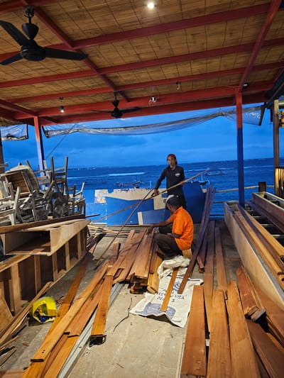 Two men are working on wooden structures on a waterfront promenade in Denpasar, Indonesia, as dusk approaches. The man on the left is wearing an orange t-shirt and dark pants and is squatting next to a pile of lumber. The man on the right is standing and wearing a dark long-sleeved shirt and a red baseball cap. He is holding a long wooden pole, appearing to direct or assist with the work. Lumber and construction materials are scattered around the two men, suggesting an ongoing project. In the background, the ocean is visible with several small boats on the water and a blue-painted structure that may be part of the promenade or a nearby building. The sky is a deep blue, indicating late evening. Above, a thatched roof is supported by red beams, with black ceiling fans installed. Wooden chairs are stacked to the left of the frame.