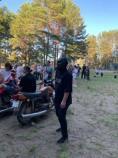 The image depicts an outdoor gathering in Chaykovsky, Russia, characterized by a sandy ground with scattered grass and a backdrop of tall pine trees under a clear blue sky. In the foreground, a person wearing a black shirt and pants, and a distinctive black skull mask with spikes, stands facing right. To the left, two vintage motorcycles are parked, with a woman seated on the one in the foreground, looking at her phone, and a man in a white t-shirt standing behind her. Other individuals are dispersed throughout the scene, some standing in groups and others seated on benches in the background. The overall atmosphere suggests a casual event or festival.