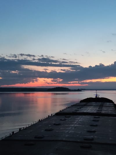 The image depicts a twilight scene over water, possibly a large lake or bay. The foreground is dominated by the dark, textured deck of a large ship or barge, running from the bottom of the frame and receding into the distance. The deck has a repetitive pattern of dark, oval shapes, likely hatches or access points.
Beyond the ship, the water reflects the colors of the sunset. The horizon line is defined by a dark band of land, possibly a forested area. Above the land, the sky displays a gradient of colors, transitioning from deep blue at the top to shades of orange, pink, and red near the horizon, where the sun is setting. Several layers of clouds are present, ranging in color from dark gray to lighter, pastel shades.
The overall lighting is dim, typical of twilight. The image does not contain any visible people. There is nothing in the image that indicates a location or association with Irdomatka, Russia.