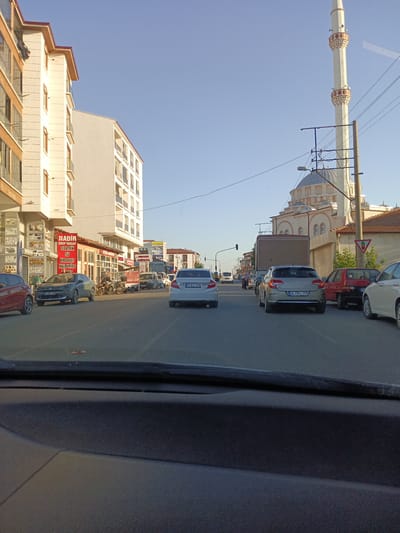 This image depicts a street scene in Bahşılı Belediyesi, Turkey, under a clear blue sky during daylight hours. On the left side of the street, a modern multi-story building with beige facade and balconies dominates the view. Adjacent to it is another white building with a similar architectural style. Several shops are visible at the ground level of the beige building, with signs indicating businesses like "NADİR GRUP İNŞAAT" and "ELEKTRIK".
The street is moderately busy with vehicular traffic. Several cars are parked and moving. A grey sedan with license plate "71 ADS 284" is parked in the foreground on the left. Further down the street, a white sedan with license plate "71 EZ 596" is visible, as is another grey sedan with license plate "34 CRU 119". A red car is parked on the right side of the road. A truck with a beige cargo box is also present.
In the background, a prominent mosque with a tall, slender minaret and a dome is visible, featuring intricate architectural details and embellishments. Several power lines and utility poles are also present, crossing the sky. The overall environment suggests a commercial and residential area within a Turkish town or city. The image appears to be taken from the interior of a vehicle, looking forward through the windshield.