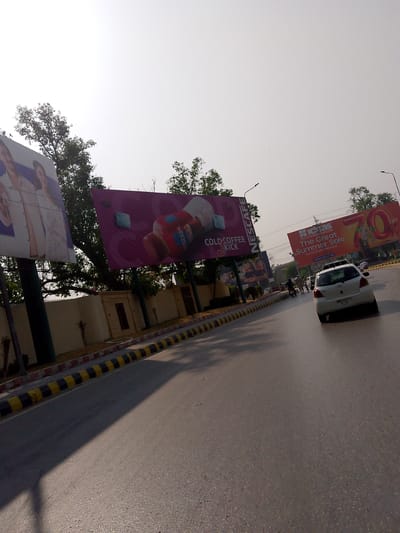 The image depicts a street scene in Peshawar City Tehsil, Pakistan, under a bright, hazy sky. A white car is visible in the foreground, driving on the right side of a paved road. The road has a distinct yellow and black striped curb. Alongside the road, there is a low, cream-colored wall.
Dominating the background are three large billboards. The leftmost billboard shows a close-up of two individuals. The central billboard features a large bottle of Nescafe Cold Coffee with the text "COLD COFFEE KICK" and "NESCAFÉ". The rightmost billboard advertises "iCASH THE Great Summer Sale" with "UP TO 70% OFF". A street light is visible above the central and rightmost billboards.
Several trees are scattered throughout the scene, with dense foliage. A few individuals, mostly appearing as silhouettes due to distance and lighting, are visible on the sidewalk and near the billboards. One person is riding a motorcycle. The overall impression is a commercial area within the city.