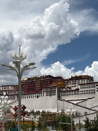 The image displays a majestic daytime view of the Potala Palace in Chengguan District, China, under a bright, partly cloudy sky. The imposing palace, built on a hill, is the central subject, showcasing its iconic red and white sections with numerous small, dark windows. Glimmers of golden roofs are visible on the upper structures of the palace.
In the foreground, a tall, decorative street lamp adorned with white, flower-like lights and intricate metalwork extends upwards from the left. Further to the right, red ornamental lanterns hang from another pole, adding vibrant cultural detail. Green foliage and trees are scattered around the base of the palace and along the hill, providing a natural contrast to the architecture.
The sky above is a clear blue, interspersed with large, fluffy white and grey clouds. A green street sign is partially visible, displaying Chinese characters that read "琼瑶多岗路" (Qiongyao Duogang Road), indicating a specific location. A traffic sign, possibly denoting a restriction, is also partly visible. The scene captures the grandeur of the historic site on what appears to be a pleasant, sunny day. No people are discernible in the frame, emphasizing the monumental scale of the palace.