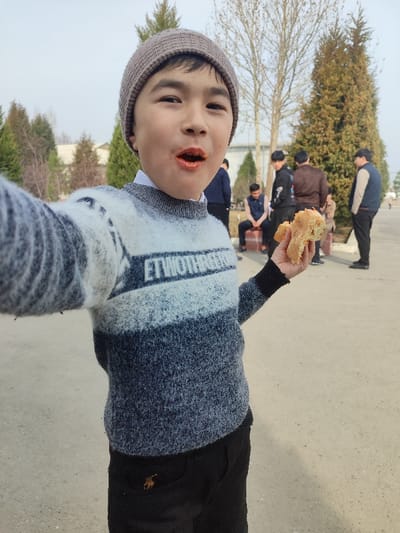 An outdoor scene in Rishtan, Uzbekistan, depicts a young boy approximately 8-12 years old as the primary subject, captured in a close-up, seemingly self-portrait style. The boy wears a grey knitted beanie, a white collared shirt, and a multi-toned knitted sweater with white, light grey, and dark grey horizontal stripes, featuring white text "ETWOTHARES" on the chest. He also wears black trousers with a small golden embroidered logo on the right hip. His mouth is open, with visible orange-red food residue around and inside it. He holds a partially consumed piece of light-colored bread or pastry in his left hand, extended towards the camera.

In the blurred background, several adult males are present. One male in a dark vest sits on a low brick structure to the right of the frame, while others stand, casually dressed in dark jackets and shirts. The ground is light-colored and unpaved. Numerous evergreen trees and deciduous trees with bare branches populate the background. A light-colored building is faintly visible in the far distance under an overcast sky. The lighting indicates natural daylight.