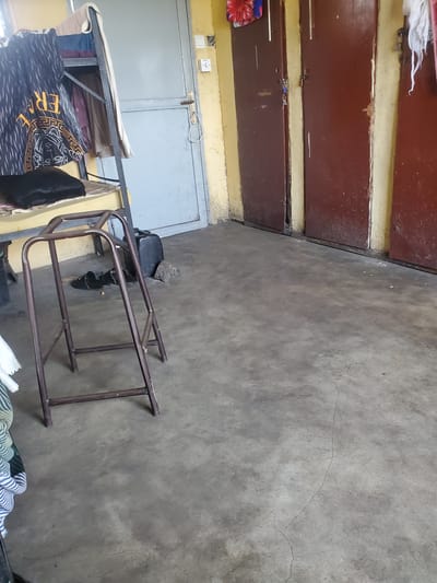 This image depicts an indoor scene, likely a room in Markurdi, Nigeria. The room appears utilitarian and somewhat cluttered. Dominating the left side of the frame are two sets of dark brown, wooden double doors, each with visible hardware and a yellow-painted trim. One set of doors is slightly ajar, revealing a glimpse of what might be a metallic surface behind it. To the right of the doors, a light grey metal door with a handle is visible. Above this door are two light-colored electrical fixtures, one resembling a light switch.
In the foreground and middle ground, a metal frame structure, possibly a cot or shelving unit, is present. It is made of brown metal tubing and appears somewhat aged. On this structure, several items are placed, including folded fabrics and what seems to be a mattress or bedding. A black garment, possibly trousers, is also visible. To the right of this structure, a small, dark seat with a stone placed on top of it is visible. A pair of black shoes are positioned near the seat.
The floor is a concrete surface, showing signs of wear and a few cracks. The overall lighting suggests an indoor environment, possibly during the daytime, though no direct light source is evident. The scene conveys a sense of everyday living in a possibly modest setting. There is no clear indication of any specific activity taking place, and the overall impression is one of stillness. The text "ER" is partially visible on a patterned fabric in the foreground.