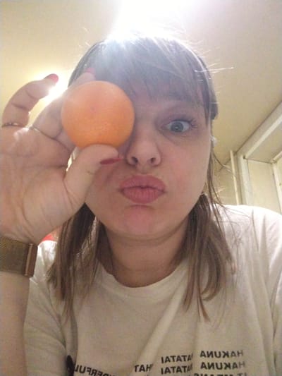A woman is taking a selfie, holding an orange over her left eye. She is pouting her lips and widening her right eye. She has brown hair with bangs and is wearing a white t-shirt with black text that includes the word "HAKUNA". She is wearing a gold watch on her left wrist and a ring on her left ring finger. The background is blurry and indistinct, appearing to be an interior space. The location in the city of Rusanovo, Russia, cannot be confirmed based on the visual information provided.