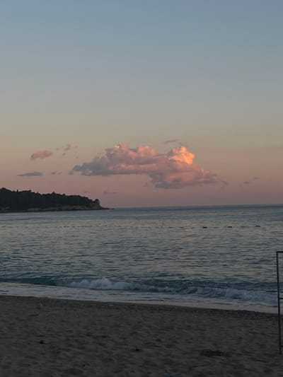 A wide shot shows a tranquil beach scene at dusk. The sky is a soft gradient of blue and pink, with a few fluffy clouds illuminated by the setting sun. In the distance, a dark, tree-covered peninsula juts out into the calm, blue sea. A line of red buoys marks a swimming area in the water. The foreground features a sandy beach with gentle waves lapping at the shore. A metal pole, possibly part of a beach umbrella or structure, is visible on the right edge of the frame. The scene is devoid of people, contributing to its serene atmosphere. The location is identified as Miljevci, Montenegro.