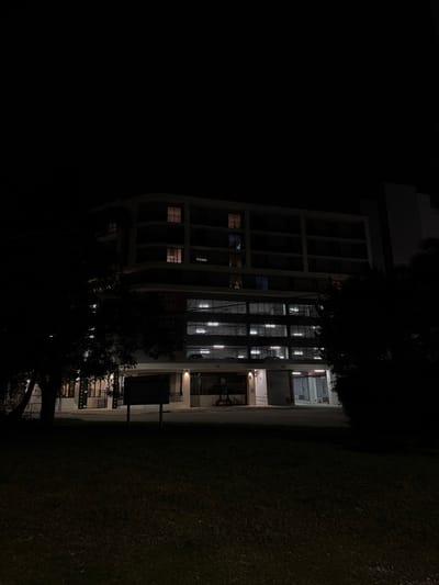 A tall, multi-story building stands illuminated at night. The building features a unique, curved exterior with numerous windows. Some windows show faint internal lighting. The building abuts a dark grassy area. A tree with dense foliage partially obscures the lower section of the building. The sky is black and unlit.