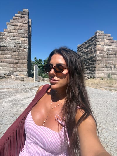 A woman is taking a selfie outdoors, with ancient stone structures and a clear blue sky in the background. She is wearing sunglasses, a pink and white gingham top, and a maroon shawl. A delicate pearl necklace adorns her neck. The sun is bright, casting shadows and highlighting her skin, suggesting it is daytime. The stone ruins, composed of large, weathered blocks, indicate a historical or archaeological site. The ground appears to be paved with cobblestones or rough stone. The overall scene evokes a sense of leisure and exploration in a warm, sunny climate. The presence of the ruins, likely in Bodrum, Turkey, places the setting in a region known for its ancient history and coastal beauty. The woman's pose and the selfie format suggest a personal moment of enjoyment and documentation of her visit.