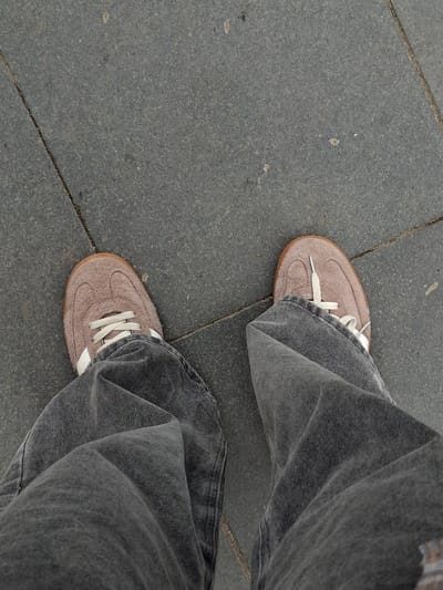 The media file shows a first-person perspective of a person standing on a paved surface. The subjects are the person's legs, clad in loose-fitting, dark grey denim jeans, and their feet, wearing light brown suede sneakers with white laces. The sneakers have a classic athletic design, possibly retro. The scene is set outdoors on a grey, textured pavement composed of large square tiles, separated by grout lines. The lighting suggests it might be daytime, with no strong shadows indicating a particular time. The weather appears neutral, as there are no elements like rain or bright sun to suggest specific conditions. The location cues are limited to the type of pavement, which is common in urban environments. No text is visible in the media file. The image is a close-up, focusing on the lower half of the person's body and their footwear. The overall impression is casual and everyday.