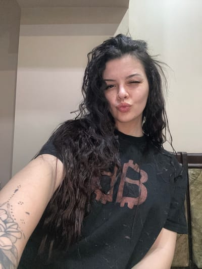 A young woman with long, dark, wavy hair, fair complexion, and freckles is depicted in a close-up, self-portrait photograph taken indoors in the city of Sliven, Bulgaria. Her left eye is closed in a wink, and her lips are pursed in a kissing gesture. A prominent tattoo, featuring floral and dotted patterns, is visible on her inner left forearm, which holds the camera. She wears a black t-shirt with a large, reddish-brown Bitcoin logo printed on the chest; the shirt is noticeably covered in lint or small debris, particularly on the right shoulder and torso. The background consists of light-colored walls with a horizontal architectural detail. A dark brown wooden furniture item with a textured, possibly green or gold, upholstered section is partially visible on the right side of the frame.