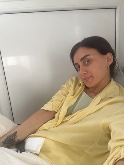 A medium shot captures a woman with dark hair and brown eyes wearing a yellow collared shirt and a light green undershirt. She has a butterfly tattoo on her left forearm. She is lying down with a white cloth and a black device next to her arm. The background is a plain white wall. The scene is indoors, and there are no immediate indications of the city of Tbilisi, Georgia.