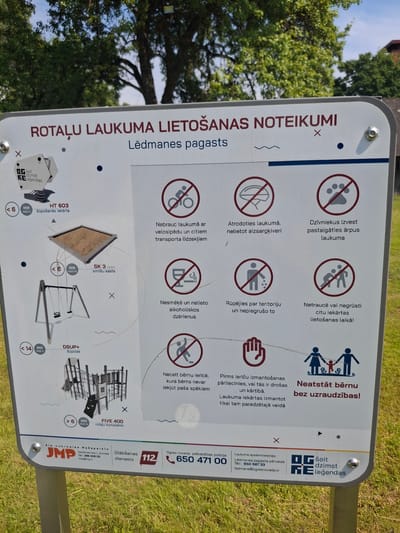 A white sign with red text and icons is mounted on a grassy area. The sign is titled "ROTĀLU LAUKUMA LIETOŠANAS NOTEIKUMI" which translates to "PLAYGROUND USAGE RULES." Below this, it specifies "Lēdmanes pagasts," indicating the location is Lēdmane parish.
The sign features various pictograms illustrating rules for playground use. These include:
- A bicycle with a line through it, indicating no bicycles.
- A helmet with a line through it, suggesting the use of protective gear.
- A paw print with a line through it, prohibiting animals.
- A cigarette and drink with lines through them, meaning no smoking or alcoholic beverages.
- A person littering with a line through it, emphasizing keeping the area clean.
- Two people with a line through them, possibly indicating no roughhousing or aggressive behavior.
- A child with a magnifying glass and a line through it, possibly meaning children should be supervised.
- A family group with a "Neatstāt bērnu bez uzraudzības!" caption, which translates to "Do not leave children unattended!"
On the left side of the sign, there are diagrams of playground equipment with age and maximum user restrictions. For example, there's a sandpit labeled "SK 3 3x3m smilšu kaste" (SK 3 3x3m sandbox) with a restriction of "< 6" and "MAX 15," and a swing set labeled "DSUP+ šūpoles" (DSUP+ swings) with "< 14" and "MAX 2." There is also a larger play structure labeled "FIVE 400 rotālu komplekss" (FIVE 400 playground complex) with "> 6" and "MAX 14."
At the bottom of the sign, there are logos and contact information for local services. This includes "SIA Jūrmalas Mežaparki" (Jūrmala Parks LLC), a phone number, an email address, "Glābšanas dienests 112" (Rescue Service 112), and information for "Ogres novada pašvaldība" (Ogre Municipality).
The background of the image shows green grass and trees, with a blue sky overhead. The sign appears to be in good condition, with some slight wear.
