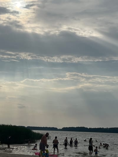 The image depicts a serene summer day at a lake in Загародскі сельскі Савет, Belarus. The sky is filled with dramatic, sun-dappled clouds, with rays of light piercing through, creating an ethereal atmosphere. In the foreground, a group of people are enjoying the water. Several individuals, including adults and children, are wading and swimming in the lake. Some are standing in the shallow water near the sandy shore, while others are further out. There are inflatable toys visible in the water. The shoreline is lined with dense green reeds and trees, forming a natural backdrop. The lake extends to the horizon, where a dark, wooded area is visible. The overall scene conveys a sense of relaxation and recreation on a beautiful summer day.
