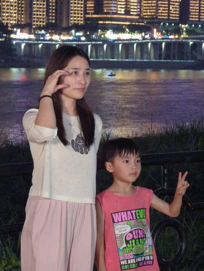 A woman and a child are posing for a photograph at night with a city skyline and a river in the background. The woman, on the left, is wearing a white long-sleeved shirt and a pink skirt. She has her hand near her face and is looking to her left. The child, on the right, is wearing a pink sleeveless shirt with graphics and is holding up two fingers in a peace sign. The background features a brightly lit city with tall buildings and a bridge spanning a river. The area where the subjects are standing appears to be an outdoor grassy area with some vegetation and a railing. The setting is likely the riverfront area of Jiangbeicheng Street, China, given the prominent cityscape.