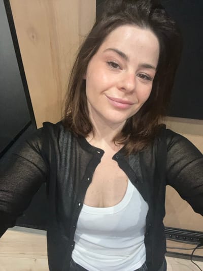A woman smiles at the camera, her face softly lit. She has fair skin, brown hair, and delicate features. She wears a white t-shirt under a sheer black cardigan. The background consists of a light wooden wall and a darker area that appears to be a corner. The image has a slight tilt, adding to a casual, self-portrait feel.