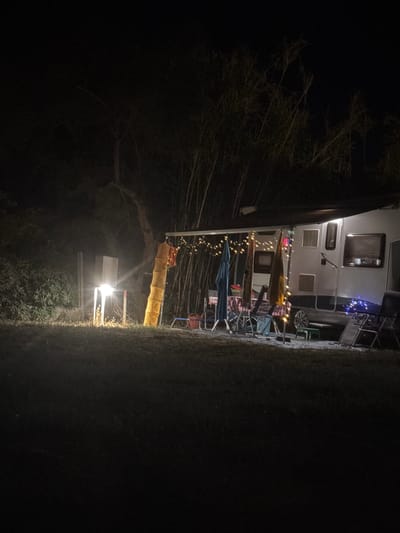 A campsite at night in Ugento, Italy, features a recreational vehicle (RV) with an awning illuminated by string lights. The RV has several windows and what appears to be a small stovetop visible through one of the windows. In front of the RV, a picnic table with chairs is set up. Two standing umbrellas are positioned near the table, one blue and one yellow. A bright light source, possibly a lamppost, is located to the left of the RV, illuminating a grassy area and some bushes. The background consists of trees and foliage, all shrouded in darkness. The scene suggests a quiet evening at a campground.