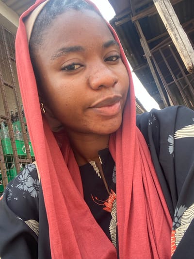 A woman wearing a red headscarf is looking at the camera. She has brown skin and is wearing a patterned top with cranes. The background is blurry but appears to be an outdoor setting with some structures visible. She has minimal jewelry. The image is angled.