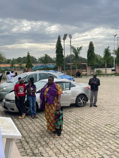 A group of people are gathered around two cars parked on a paved lot. The scene appears to be outdoors, with trees and a building in the background. The sky is cloudy, suggesting it might be overcast or late afternoon.
In the foreground, several people are standing and interacting. A woman in a vibrant, patterned outfit and a purple headscarf is at the center. To her left, two younger women are leaning against a white car, one wearing a purple t-shirt with text, and the other in a red t-shirt with a Bitcoin logo and text. A young child in a colorful garment lies on the hood of the white car, being attended to by an adult. Another man in a black jacket and cap is standing near the rear of the white car, holding something in his hand. Further back, a person in a white garment is visible near another car.
The activity appears to be a social gathering or possibly a community event. The individuals are dressed casually, and their interactions suggest a relaxed atmosphere. The presence of the cars indicates they may have arrived or are preparing to depart. The Bitcoin logo on the red t-shirt is a notable detail. The setting suggests an urban or semi-urban environment in Barakin Lerawi, Nigeria, given the architectural style of the building and the types of trees.