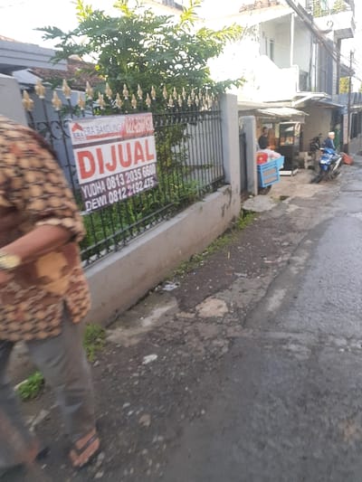 The image shows a person wearing a brown and white patterned shirt and brown pants walking on a narrow street in Cimenyan, Indonesia. The person is walking past a black metal gate with a sign that reads "Dijual" which means "for sale" in English. The sign also includes phone numbers for YUDHA and DEWI. The gate leads to a property with a white painted wall and a small house. There is a green bush in the foreground. The street is paved with asphalt but has a gravel patch. In the background, there are two people standing near a small cart. There is a motorcycle in the background. The street is narrow and appears to be in a residential area. The sky is overcast. The location is in the city of Cimenyan, Indonesia, based on the provided coordinates: -6.888 latitude and 107.652 longitude.