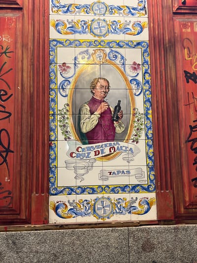 The image displays a close-up of a decorated tiled panel on the exterior of a building in Madrid, Spain. The panel features an oval portrait of a man in traditional attire, holding a bottle of wine and a glass. Below the portrait, the text "CERVECERIA CRUZ DE MALTA" is inscribed, followed by the word "TAPAS" on a banner. The tiled panel is framed with intricate blue and yellow floral motifs, and at the top and bottom, there are depictions of mythical creatures and crosses. The tiled panel is set within a weathered wooden door that has been graffitied with red and black spray paint. The door is flanked by other wooden elements and sits above a granite-like base. The overall impression is of an old-world establishment in a vibrant urban setting.