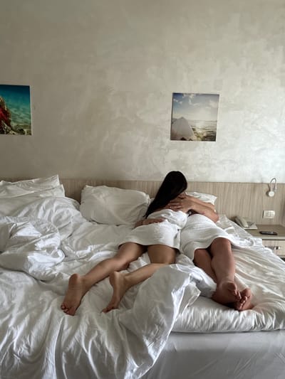 The image captures two individuals, likely a couple, intimately resting in a bed. They are partially covered by white bath towels and surrounded by crumpled white bed sheets and pillows, suggesting a relaxed or morning setting. The person on the left has long, dark hair, with their face obscured by their arm and hair as they embrace or lie close to the other person. The second individual's legs and feet, also wrapped in a white towel, are visible and intertwined with the first person's, conveying a sense of closeness and comfort.

The setting appears to be a bedroom, possibly in a hotel, given the functional wall-mounted features. The wall to the left features a subtly textured, light gray finish. Two framed pictures adorn this wall: the upper one depicts a surfboard in clear water under a bright, cloudy sky, while the lower one showcases a vibrant underwater coral reef. Above the light-colored wooden headboard, a white wall-mounted light fixture, an electrical outlet, and a white landline phone are visible, further reinforcing a hotel environment.

The overall atmosphere is tranquil and intimate, emphasizing rest and connection. The framed pictures hint at a tropical or coastal location, consistent with the provided location context of Safaga, Egypt. No discernible text is visible in the image.