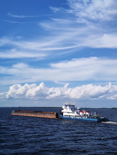 A barge train, consisting of a tugboat and a long, flat barge, moves across dark blue, choppy water under a bright blue sky filled with white, wispy clouds and larger cumulus formations. The tugboat, primarily white with blue accents and red detailing, has multiple decks and appears to be in motion, creating a wake behind it. The barge, a long, rectangular vessel, is laden with what appears to be dark material, possibly cargo, and shows signs of weathering. The horizon line is visible, with a dark, forested shoreline in the distance, suggesting a river or a large lake in the city of Maksakovo, Russia. The overall lighting indicates it is daytime, likely mid-day, with the sun positioned to illuminate the scene.